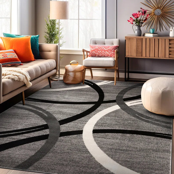 A modern living room with a Devonshire Abstract Gray/Black/Ivory Area Rug, beige sofa, colorful pillows, wooden sideboard, and a white pouf.