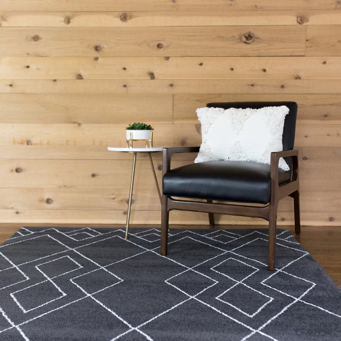 A cozy corner with a black armchair, white pillow, and small round table featuring a plant on the Dedham Geometric Black Area Rug.