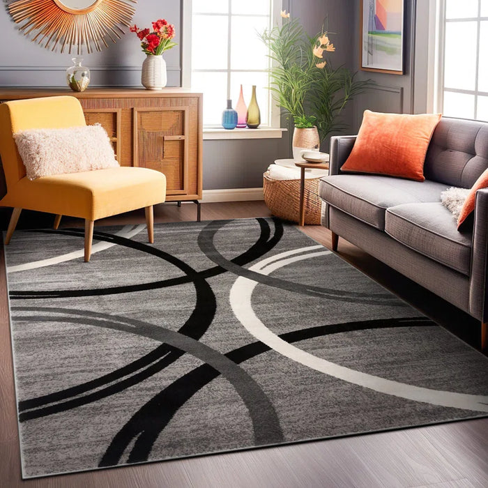 Modern living room with a Devonshire Abstract Gray/Black/Ivory Area Rug, yellow armchair, gray sofa with orange pillows, and decorative plants near the windows.