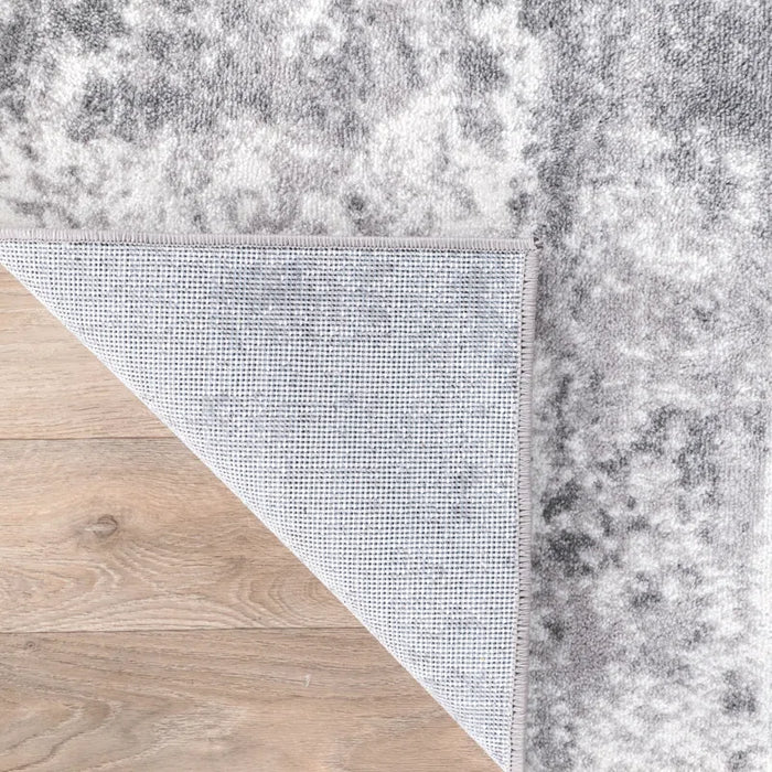 A corner of the Willington Abstract Ivory/Dark and Light Gray Area Rug is flipped over on a light wooden floor, showing its underside.