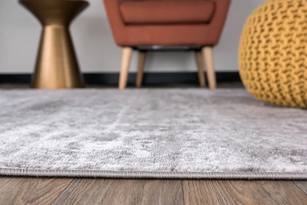 Close-up of the Willington Abstract Ivory/Dark and Light Gray Area Rug on a wooden floor, with a blurred brown chair, round gold table, and yellow pouf in the background.