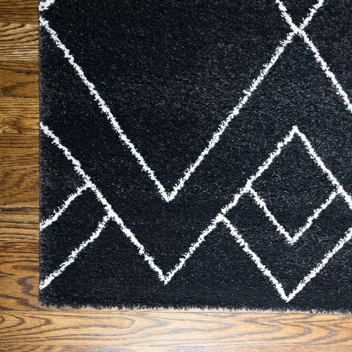The Dedham Geometric Black Area Rug, featuring intersecting white geometric lines, is placed on a dark wooden floor with only a corner visible.