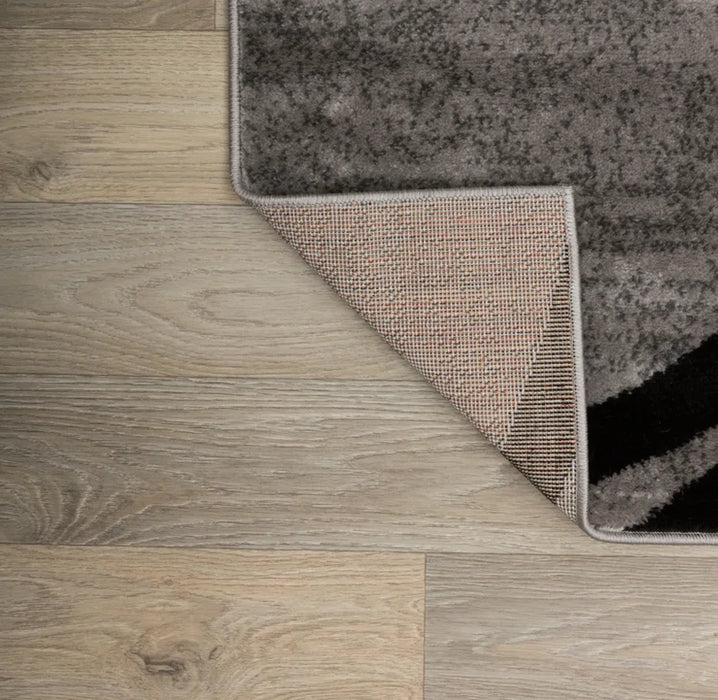 A corner of the Devonshire Abstract Gray/Black/Ivory Area Rug is folded over, revealing its underside, displayed on a light wood floor.