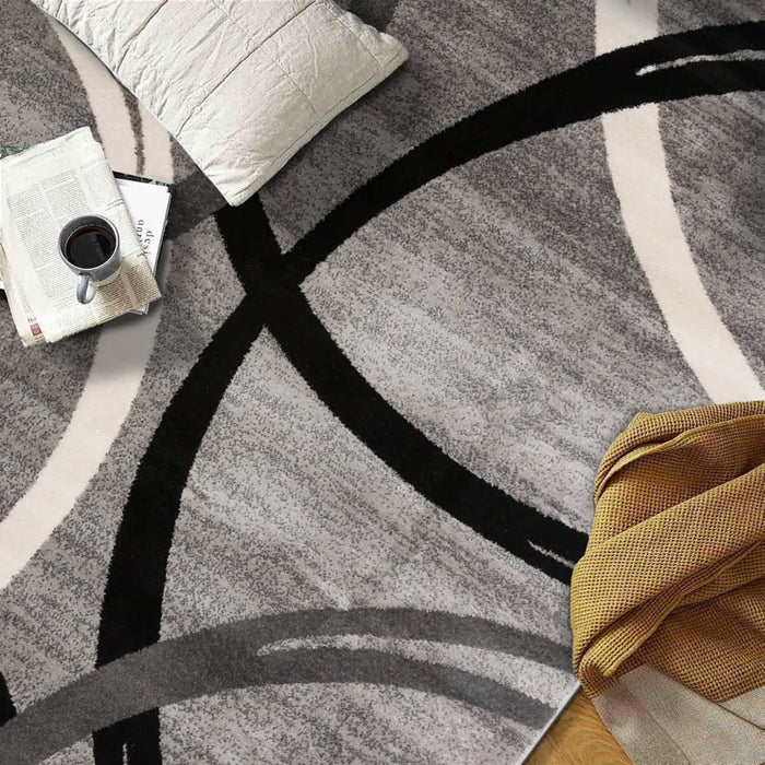 Devonshire Abstract Gray/Black/Ivory Area Rug with a pillow, newspaper, cup of coffee, and a blanket on a wooden floor.