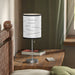 The Random Fact Table Lamp sits on a wooden nightstand by a bed with books and a remote, near green-curtained window.