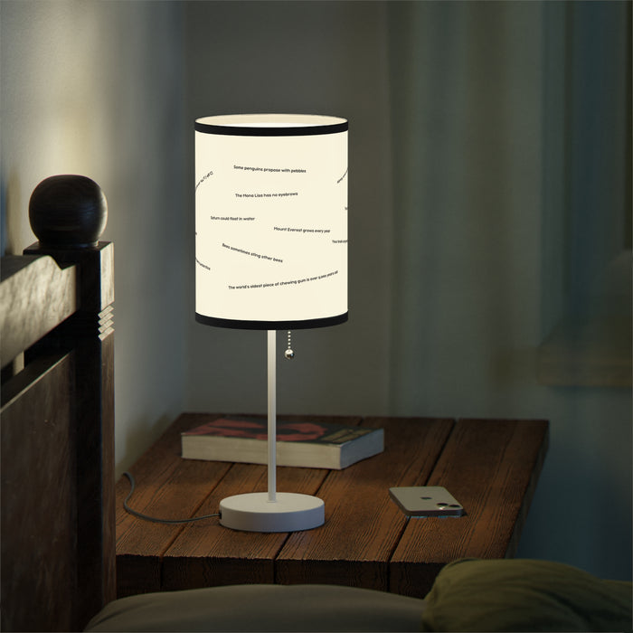 A Random Fact Table Lamp glows on a bedside table with a book and smartphone beside a bed.