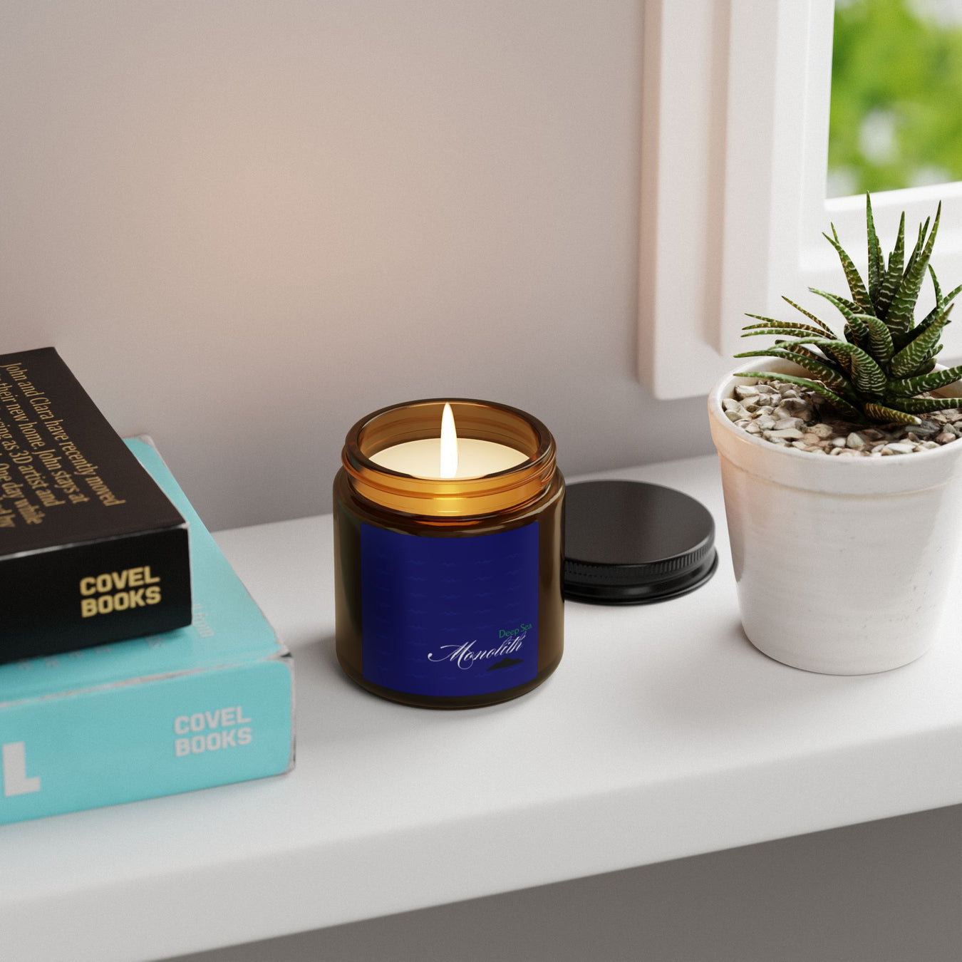 An Atlantis Whisper candle from the Deep Sea Monolith Collection sits on a windowsill with books and a potted succulent.