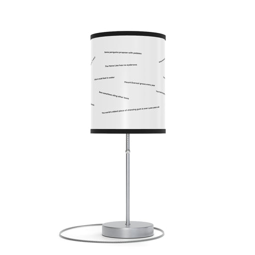 The Random Fact Table Lamp has a sleek metallic base and a modern cylindrical shade with scattered text phrases.