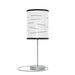 The Random Fact Table Lamp has a sleek metallic base and a modern cylindrical shade with scattered text phrases.