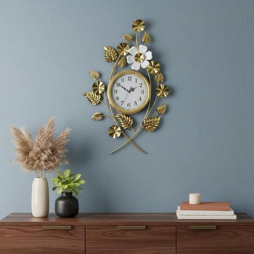 Decorative wall clock with floral design on a blue wall above a wooden shelf with plants and books.