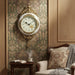 Decorative wall clock on a wall with ornate wallpaper and a chair.