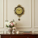 Decorative wall clock with floral arrangements and books on a wooden cabinet against a beige wall.