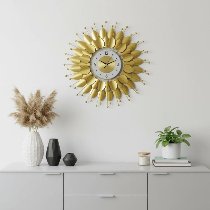 Decorative gold wall clock on a white wall with a shelf below featuring vases and plants.