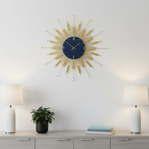Decorative wall clock with gold rays on a white wall above a wooden cabinet with lamps and a plant.