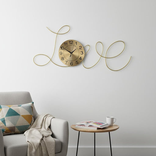 Gold wall clock with 'LOVE' design on a white wall above a gray sofa with a geometric pillow.