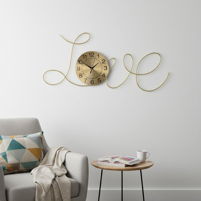 Gold wall clock with 'LOVE' design on a white wall above a gray sofa with a geometric pillow.