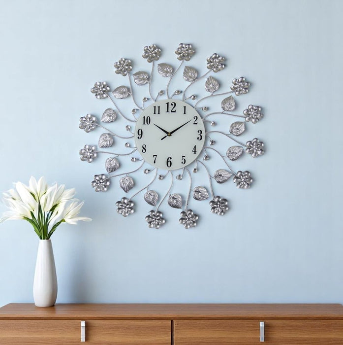 Decorative wall clock with floral design on a light blue wall, next to a wooden cabinet and white vase with flowers.