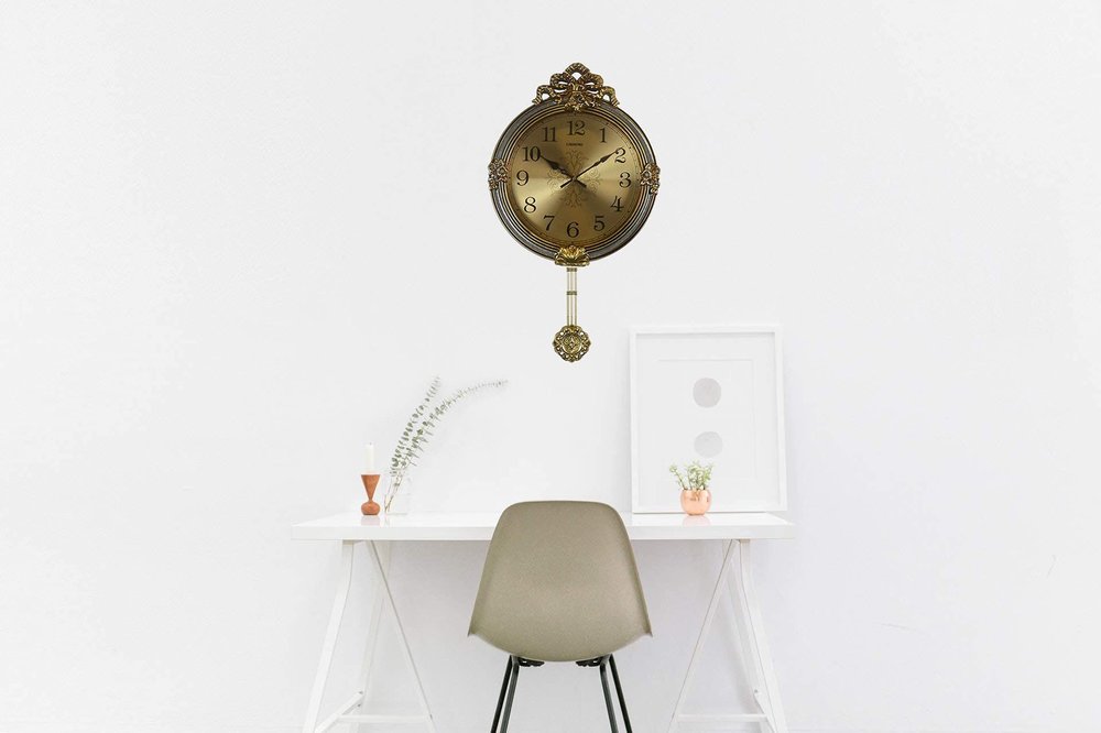 Decorative wall clock with a pendulum on a white wall above a table with a chair.