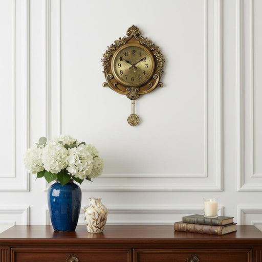 Decorative wall clock with a vase of flowers and books on a wooden table against a white paneled wall.