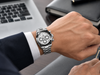 Hand wearing a silver watch with a blurred background of a laptop and notebook.